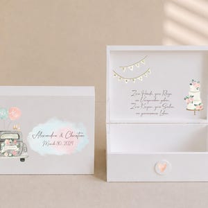 May include: A white wooden keepsake box featuring a watercolor illustration of a vintage car with balloons, the names Alexandra & Christian, and the date March 30, 2024. The open lid displays a wedding cake, bunting, and German text.
