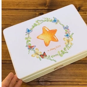 K&ouml;nnte beinhalten: Eine wei&szlig;e Holzkiste mit einem bemalten Design eines gelben Sterns, umgeben von gr&uuml;nen Bl&auml;ttern und bunten Schmetterlingen. Der Text "4. M&auml;rz 2021" ist auf die Kiste gedruckt.