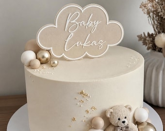 Könnte beinhalten: Ein runder, cremefarbener Kuchen mit einem beigen, wolkenförmigen Tortenstecker, der den weißen Schriftzug Baby Lukas trägt. Der Kuchen ist mit einem kleinen, strukturierten Teddybären, matten weißen und goldfarbenen Kugeln sowie Blattgold verziert.