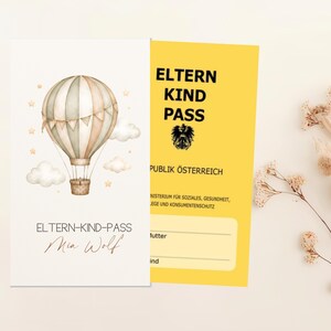 May include: A white card featuring a watercolor hot air balloon with clouds and stars, labeled ELTERN-KIND-PASS and Mia Wolf. Beside it, a yellow card titled ELTERN KIND PASS REPUBLIK &Ouml;STERREICH with an eagle emblem and blank lines for names.