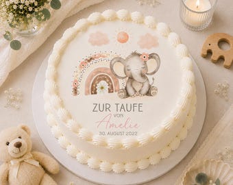 May include: A round white cake decorated with a watercolor illustration of a gray elephant, rainbow, and sun. The cake has a piped buttercream border and text that reads "ZUR TAUFE VON Amelie 30. AUGUST 2022". A teddy bear and candle are in the background.