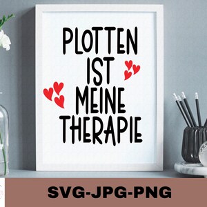 Puede incluir: Impresión enmarcada en blanco con texto negro que dice "Plotten ist meine Therapie" con tres corazones rojos sobre el texto. La impresión está sobre un fondo gris con un jarrón de flores blancas y un porta lápices negro sobre una mesa.