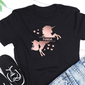 May include: Black t-shirt with a rose gold unicorn silhouette and the name "Maria" printed on it. The unicorn has a horn, mane, and tail. There are stars and hearts around the unicorn.