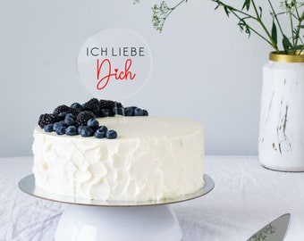 May include: White cake with white frosting and blueberries and blackberries on top. A clear acrylic cake topper with the text "ICH LIEBE Dich" in black and red.