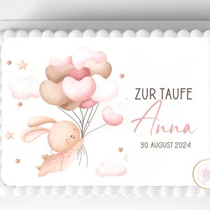 May include: A white cake with a pink and brown bunny holding pink and brown balloons. The cake is decorated with the text "Zur Taufe Anna 30. August 2024".