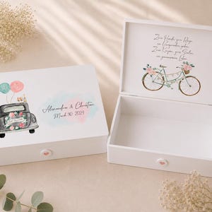 May include: Two white wooden keepsake boxes with hinged lids. The closed box features a watercolor illustration of a car with balloons and the text Alexandra & Christian March 30, 2024. The open box shows a bicycle illustration and German text.