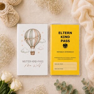 May include: Two rectangular booklets, one white with a watercolor hot air balloon illustration and the text "MUTTER-KIND-PASS Mia Wolf," and one yellow with the black text "ELTERN KIND PASS REPUBLIK &Ouml;STERREICH" and blank lines for names.