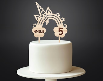 Op de afbeelding: Houten taarttopper met een regenboog met een eenhoornhoorn en sterren, met de naam "AMELIE" en het nummer "5" op wolken.