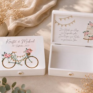 May include: Two white wooden keepsake boxes. One box is closed, featuring a bicycle illustration with pink flowers, the names Kerstin & Michael, and the date 30. September 2023. The open box shows German text, a wedding cake, and bunting.