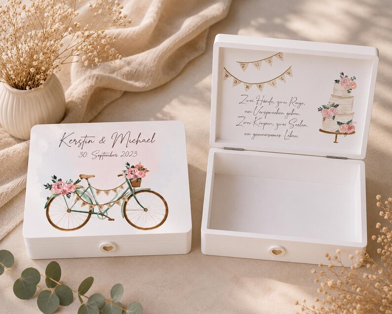 Pu&ograve; includere: Due scatole ricordo in legno bianco. Una scatola &egrave; chiusa e presenta un'illustrazione di bicicletta con fiori rosa, i nomi Kerstin & Michael e la data 30. September 2023. La scatola aperta mostra testo in tedesco, una torta nuziale e festoni.