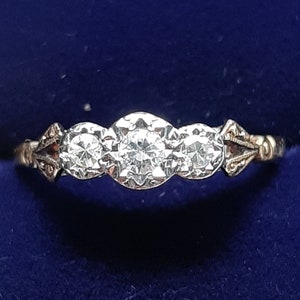 May include: A gold ring with three round, clear diamonds set in a row. The ring is in a blue velvet jewelry box. The ring has decorative filigree details on the sides.
