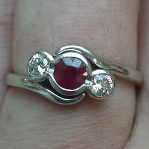 May include: A silver ring with a red ruby and two smaller diamonds. The ring is on a finger.