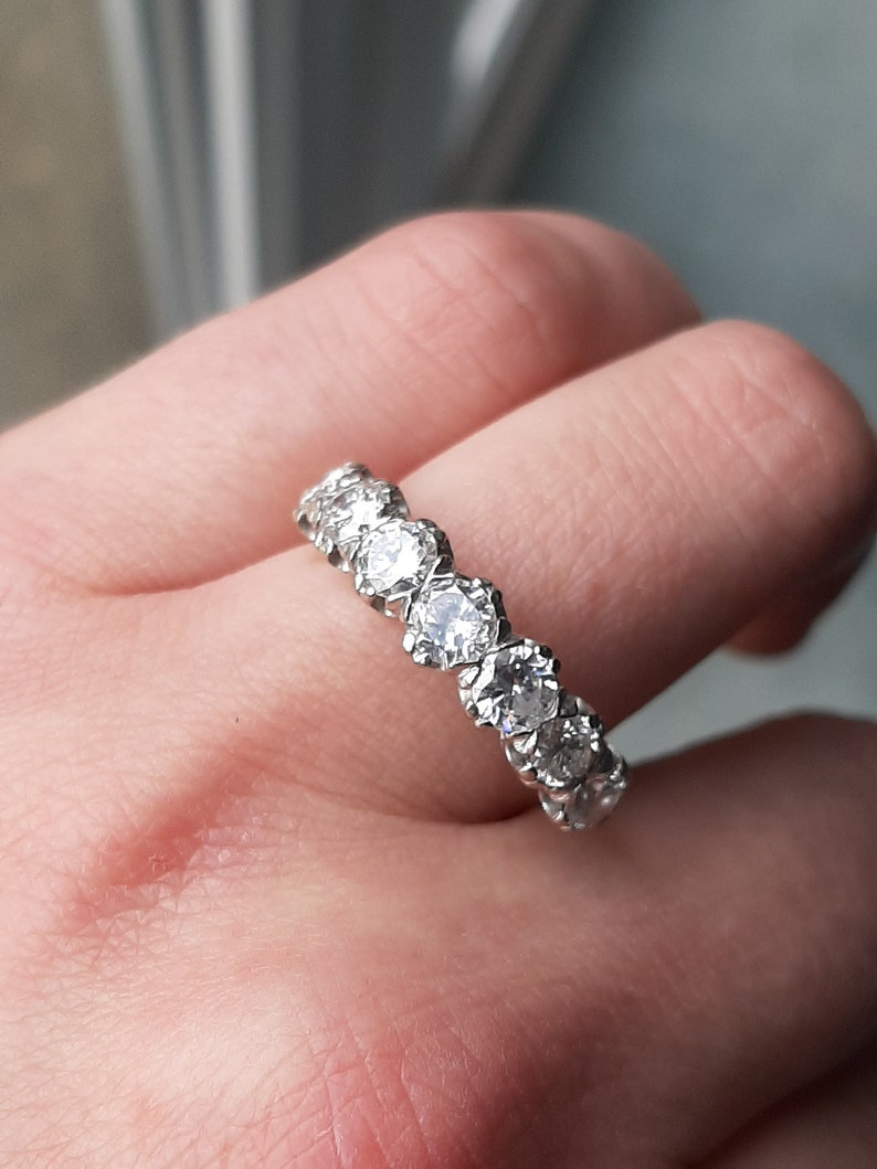May include: A silver band with a row of round, clear gemstones set in a prong setting.