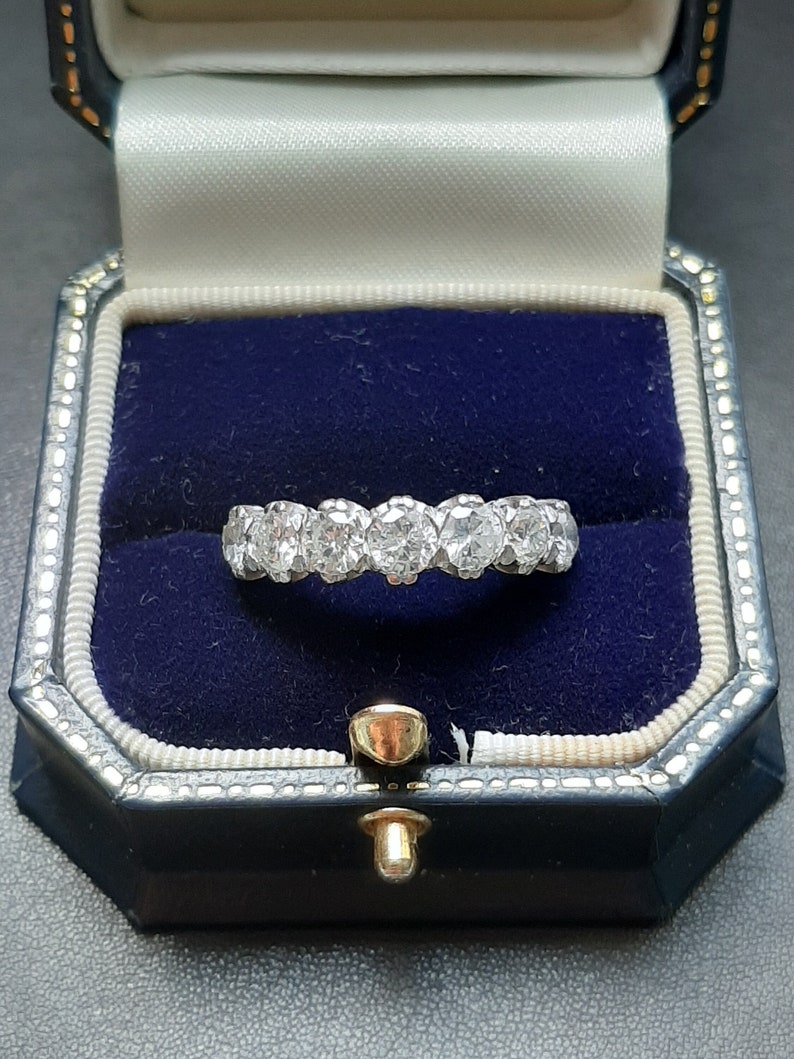 May include: A diamond eternity band ring with a row of round brilliant diamonds set in a white metal band. The ring is displayed in a blue velvet ring box.
