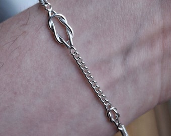 May include: A silver bracelet featuring a knot design and a chain link style. The bracelet has a clasp fastening.