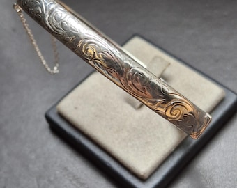 Könnte beinhalten: Ein silberfarbenes, graviertes Armreifarmband mit einem verzierten Blumenmuster. Das Armband ist rechteckig und hat eine zarte Kette. Das Design ist detailliert und scheint Vintage oder antik zu sein.