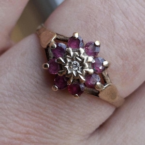 May include: A gold-toned ring featuring a floral design. The ring's center showcases a small, sparkling diamond surrounded by a cluster of deep red gemstones. The band has a delicate, openwork design.