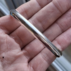 Op de afbeelding: Een gepolijste zilverkleurige metalen armband met een glad, afgerond en reflecterend oppervlak. De armband heeft een eenvoudig, doorlopend en solide ontwerp zonder zichtbare patronen, gravures of extra decoratieve elementen.