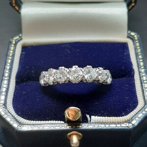 May include: A diamond eternity band ring with a row of round brilliant diamonds set in a white metal band. The ring is displayed in a blue velvet ring box.