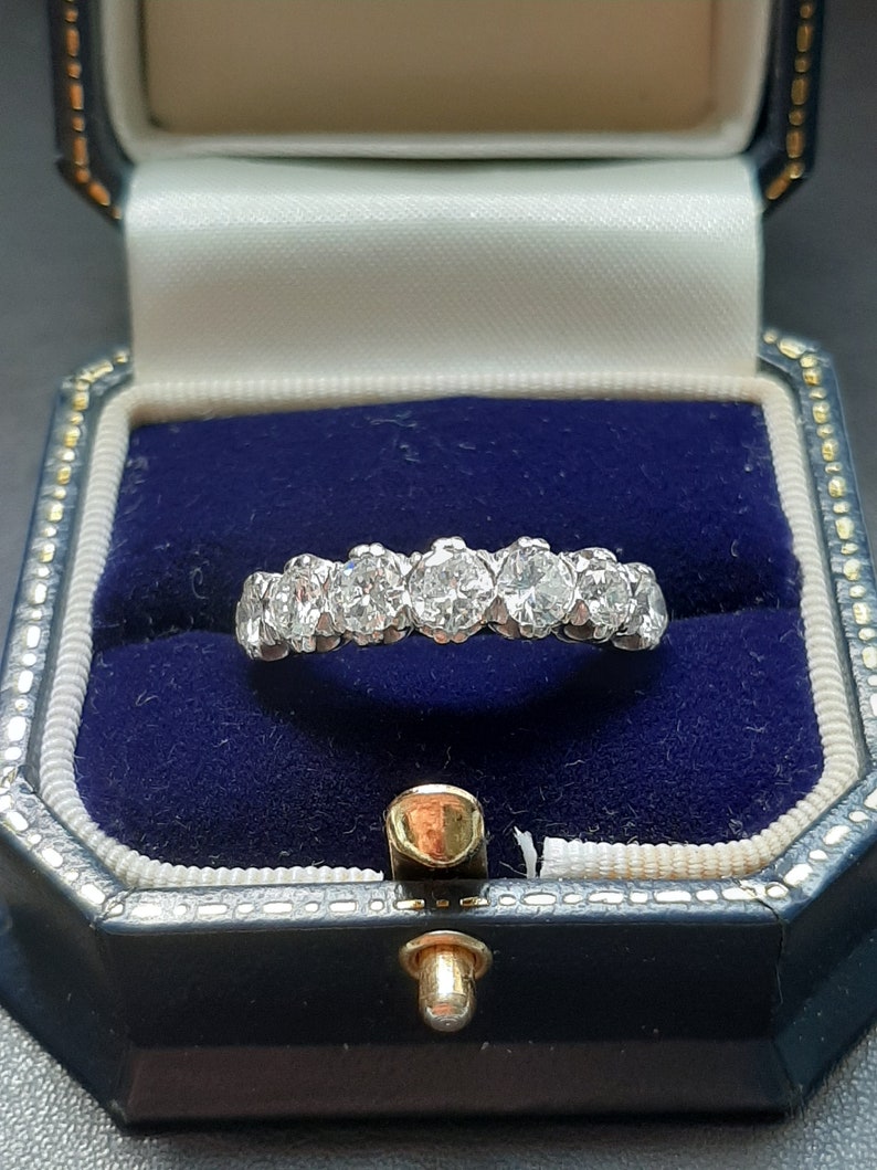 May include: A diamond eternity band ring with a row of round brilliant diamonds set in a white metal band. The ring is displayed in a blue velvet ring box.