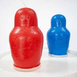 以下が含まれることがあります： 顔と花柄が描かれた赤と青のプラスチック製の入れ子人形2体。人形は白い台座の上に置かれています。
