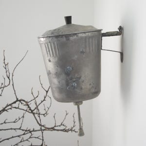 Könnte beinhalten: Ein wandmontiertes Metallbecken mit verwitterter grauer Oberfläche, ausgestattet mit einem Deckel mit kleinem schwarzem Knopf, vertikalen Rillen am oberen Rand und einem zentralen, nach unten gerichteten Metallauslauf.