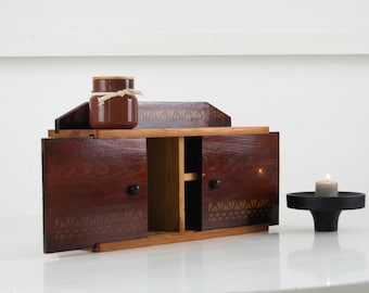 May include: A wooden cabinet with two doors, open to reveal a light-colored interior. A small brown jar with a lid and tied with twine sits on top. A black candle holder with a lit candle is to the right.