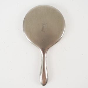 May include: A silver-toned hand mirror with a round head and a long, curved handle. The mirror's surface has a subtle sheen, and an ornate letter "F" is engraved on the back. The handle is smooth and rounded, providing a comfortable grip.