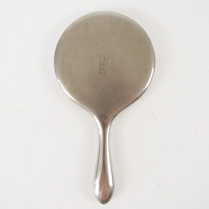 May include: A silver-toned hand mirror with a round head and a long, curved handle. The mirror's surface has a subtle sheen, and an ornate letter "F" is engraved on the back. The handle is smooth and rounded, providing a comfortable grip.