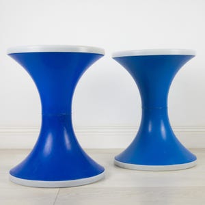 May include: Two blue, hourglass-shaped stools with white trim. The stools have a wide base and top, connected by a narrower midsection. The stools are made of a glossy material and are placed on a light-coloured floor.