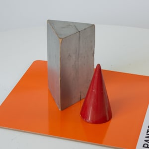 May include: A gray, triangular prism and a red cone rest on an orange surface. The prism is made of wood and has a weathered appearance. The cone is smooth and glossy. The objects are set against a white background.