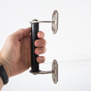 May include: An antique door handle with a black cylindrical grip and two silver-toned mounting plates. The handle is held against a white background, showcasing its vintage design and metal construction.