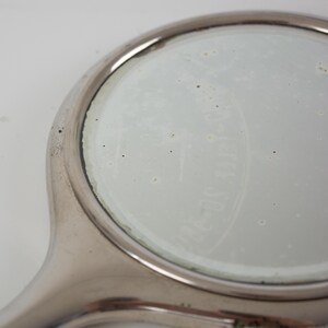 May include: A close-up of a vintage, round, hand-held mirror with a silver-colored frame and handle. The mirror's surface is slightly cloudy, and faint text is visible on the glass. The handle curves smoothly into the circular mirror.