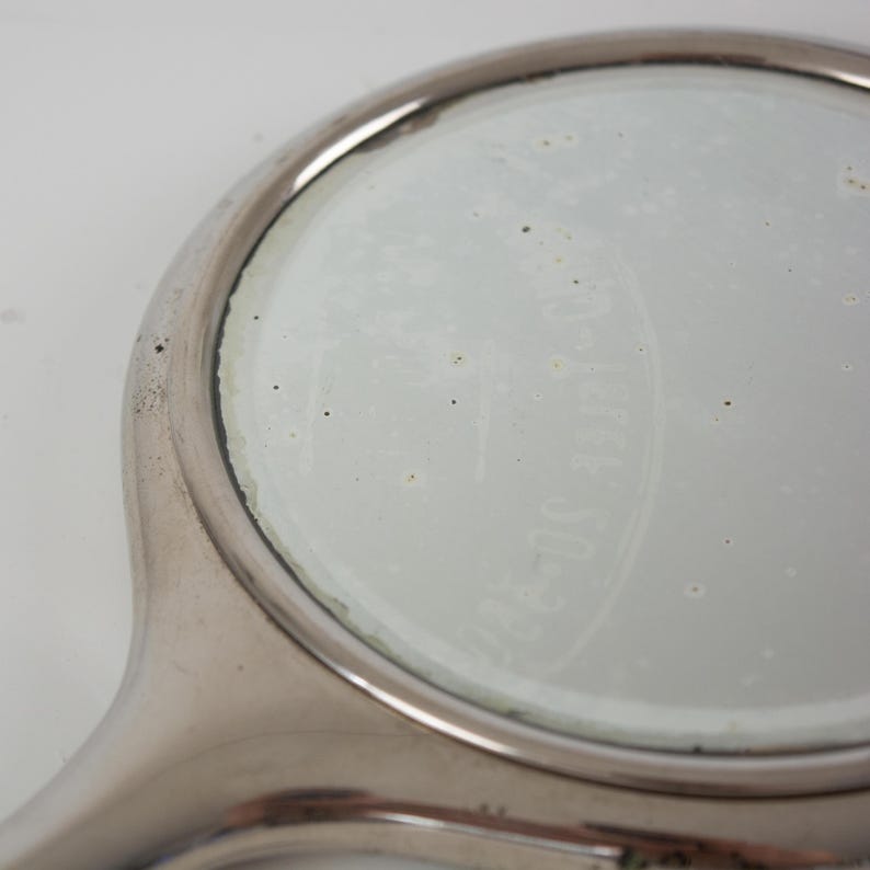 May include: A close-up of a vintage, round, hand-held mirror with a silver-colored frame and handle. The mirror's surface is slightly cloudy, and faint text is visible on the glass. The handle curves smoothly into the circular mirror.