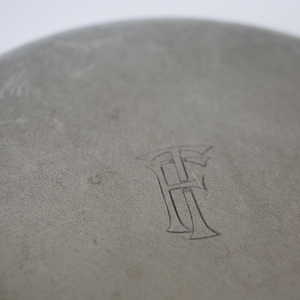 May include: Close-up of a gray, circular object with a textured surface. The object has an engraved monogram of two stylized letters, possibly "H" and "F". The background is a soft, neutral tone.
