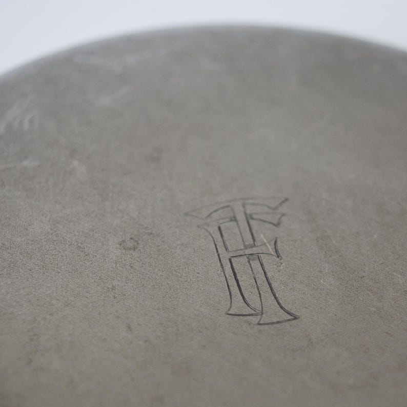 May include: Close-up of a gray, circular object with a textured surface. The object has an engraved monogram of two stylized letters, possibly "H" and "F". The background is a soft, neutral tone.