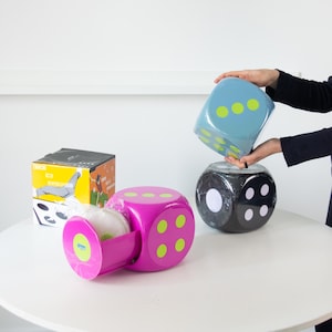 May include: A selection of large dice in shades of blue, black, and pink, each marked with green dots. One die is held up, while another is open, showing a white item within. A box with illustrations is also visible.