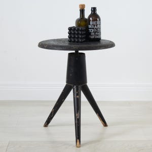 May include: A black tripod side table with a round top and distressed finish. On the table sits a black cube with a textured surface, a small amber glass bottle with a cork stopper, and a dark brown glass bottle with the text BIERE DES AMIS 33CL.