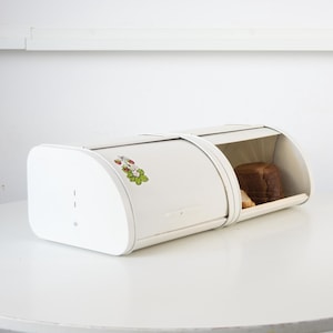 Op de afbeelding: Een witte metalen broodtrommel met een roldeksel. De broodtrommel heeft een aardbei- en bladsticker aan de zijkant. Het deksel is open en onthult een brood in de broodtrommel. De broodtrommel staat op een wit oppervlak.