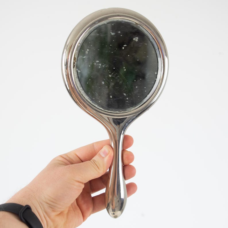 May include: A vintage hand mirror with a silver-colored frame and handle. The round mirror has a dark, reflective surface. The handle is elongated and curved, with a smooth, polished finish. The mirror is held in a hand against a white background.
