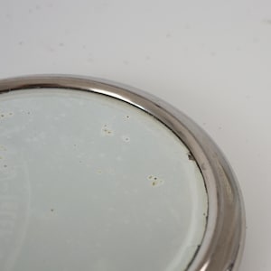 May include: A round mirror with a silver metal frame. The mirror's surface is slightly speckled, and the frame has a polished finish. The image is a close-up of the mirror's edge and surface.