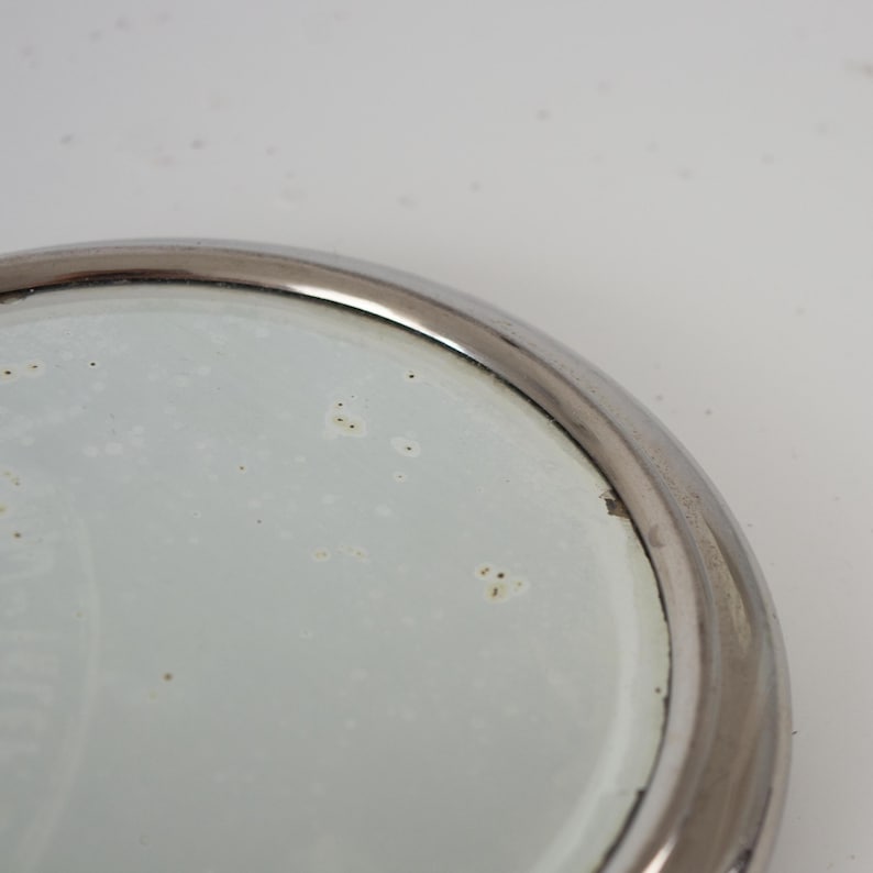 May include: A round mirror with a silver metal frame. The mirror's surface is slightly speckled, and the frame has a polished finish. The image is a close-up of the mirror's edge and surface.