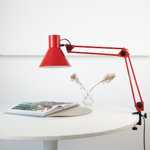 May include: A red desk lamp with an adjustable arm and conical shade is clamped to a white table. A book and a small glass vase with dried plants are on the table. The lamp's arm extends approximately 61 cm.