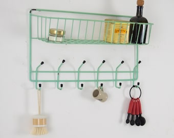 May include: A mint green metal wall-mounted organizer with a wire basket shelf and a row of hooks. The shelf holds a jar, a tea box, and a bottle. A brush, a mug, and measuring spoons hang from the hooks.