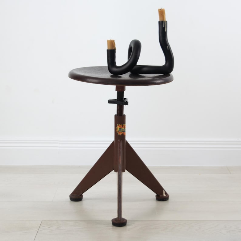 K&ouml;nnte beinhalten: Ein brauner, verstellbarer Hocker mit runder Sitzfl&auml;che und dreibeinigem Fu&szlig;, auf dem ein schwarzer, geschwungener, r&ouml;hrenf&ouml;rmiger Kerzenhalter steht. Der Halter enth&auml;lt zwei helle Stabkerzen. Am Mittelpfosten befindet sich ein kleines, buntes Etikett.
