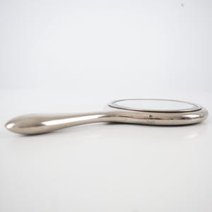 May include: A silver-toned hand mirror with a rounded oval mirror and a long, curved handle. The mirror has a polished, reflective surface and a sleek, modern design. The handle is smooth and ergonomically shaped for comfortable use.