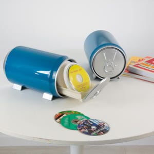Könnte beinhalten: Zwei blaue zylindrische Aufbewahrungsbehälter in Form von Getränkedosen mit silbernen Deckeln mit Zuglasche. Ein Behälter ist geöffnet und zeigt ein weißes Innenfach mit mehreren Compact Discs, darunter eine mit einem gelben Etikett.