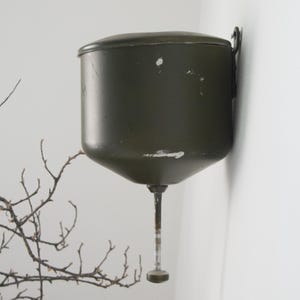 May include: A wall-mounted olive green metal container with a rounded top and a tapered bottom. A thin, vertical metal rod extends downward from the center of the base, ending in a small circular knob. The surface shows minor paint wear and scratches.