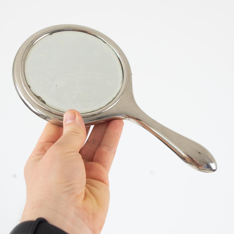 May include: A silver-toned hand mirror with a round reflective surface and a long, curved handle. The mirror is held in a hand against a white background. The mirror's frame and handle have a shiny finish.