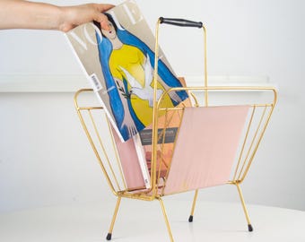 Puede incluir: Un revistero de metal dorado con un asa negra y paneles de tela rosa. El revistero contiene varias revistas, incluida una con una mujer con un vestido amarillo en la portada. El revistero tiene un diseño moderno de mediados de siglo.